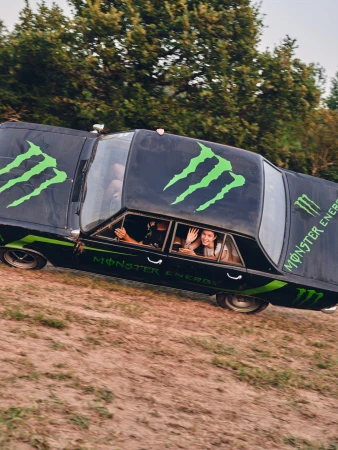 An modified black car with Monster Energy branding drives along a dirt road with two people waving from the windows, trees in the background.