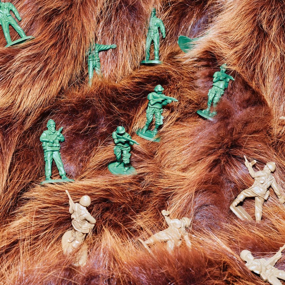 Overhead shot of green and tan plastic toy soldiers in various poses arranged on textured brown faux fur, creating a miniature battlefield scene.
