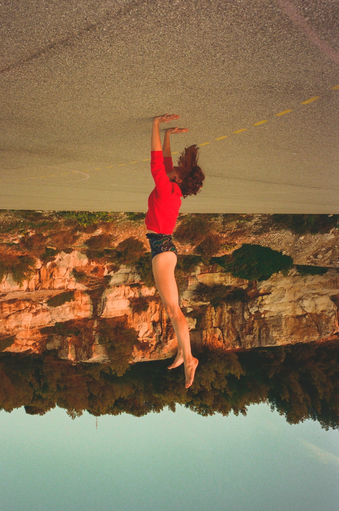 Woman in red shirt poses athletically outdoors against a cliffside backdrop, shot with artistic post-processing and altered color tones.