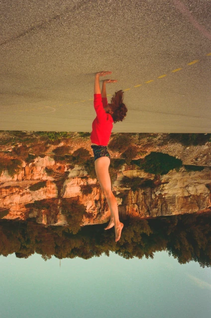 Woman in red shirt poses athletically outdoors against a cliffside backdrop, shot with artistic post-processing and altered color tones.