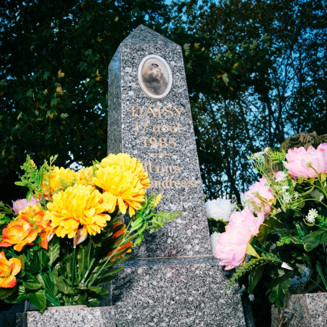 Granite headstone in outdoor cemetery featuring a portrait, text inscription, and surrounded by floral arrangements with a tree backdrop.