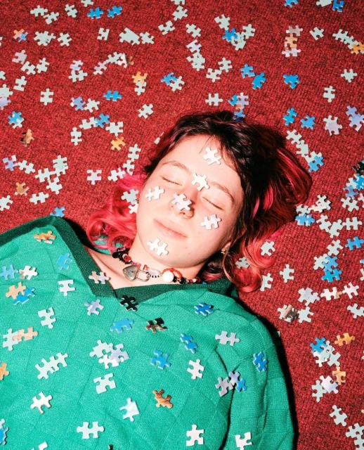 Woman with pink hair lies on a red carpet covered in jigsaw puzzle pieces, some scattered on her face, wearing a green sweater.