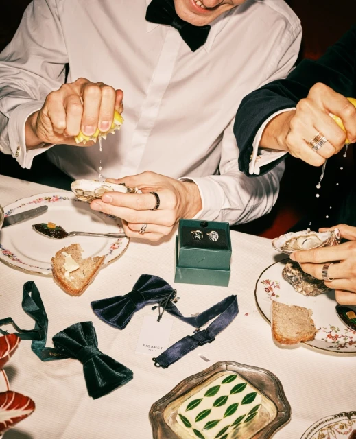 Two men squeeze lemons over oysters at a formal dining table set with bow ties, plates, silverware, and drinks for an elegant meal.