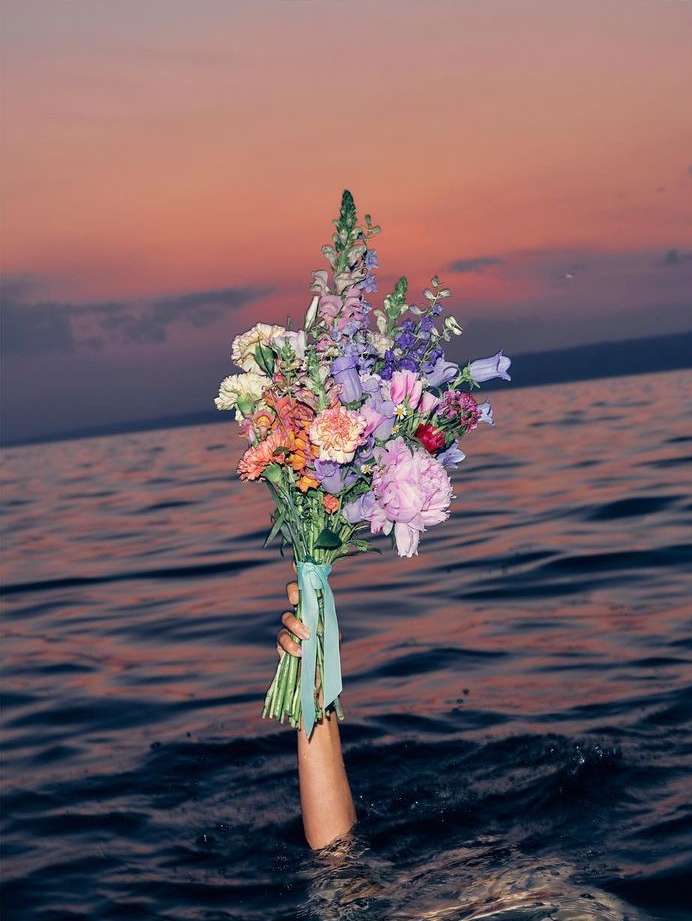 Arm holding a colorful flower bouquet rising from the water at sunset. Ocean with horizon line, orange and pink sky. Floral arrangement.
