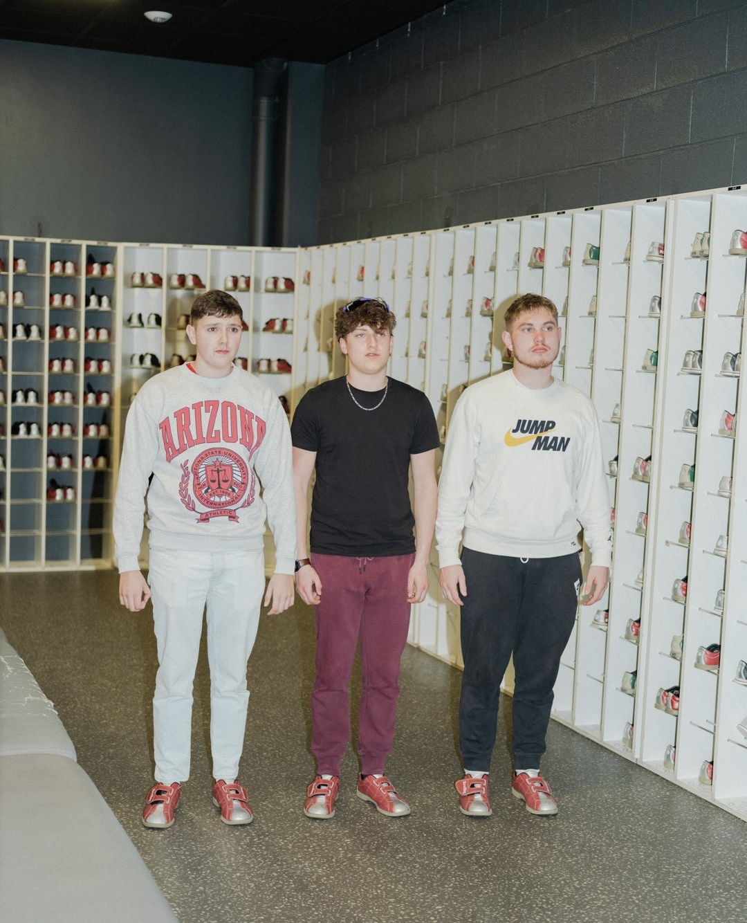 Three young adult men pose indoors in front of shelves of shoes, wearing sweatshirts and casual pants with red-and-silver sneakers.
