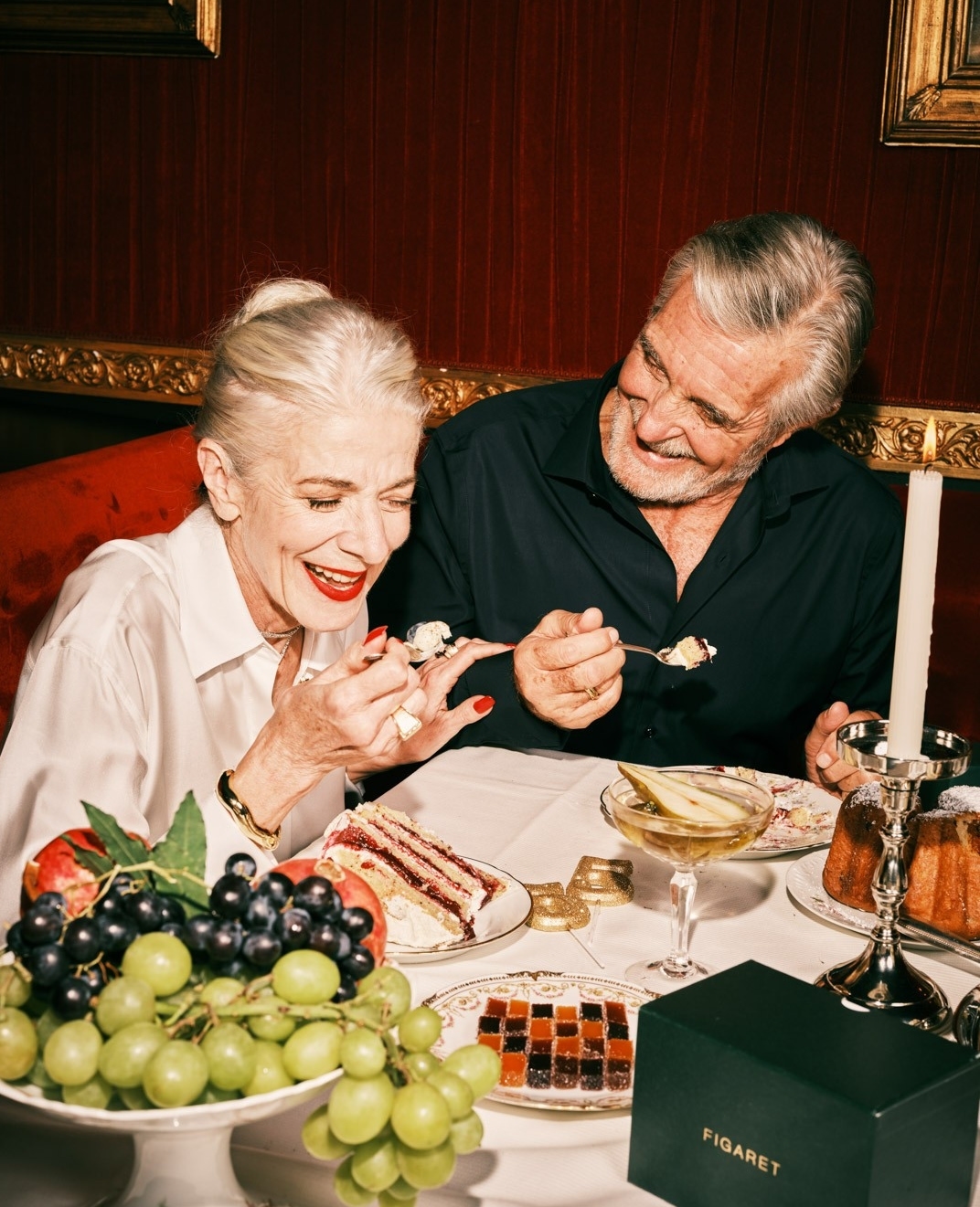 Older couple laughing while sharing dessert and cocktails in a dimly lit restaurant. Anniversary dinner with cake, fruit, and drinks.
