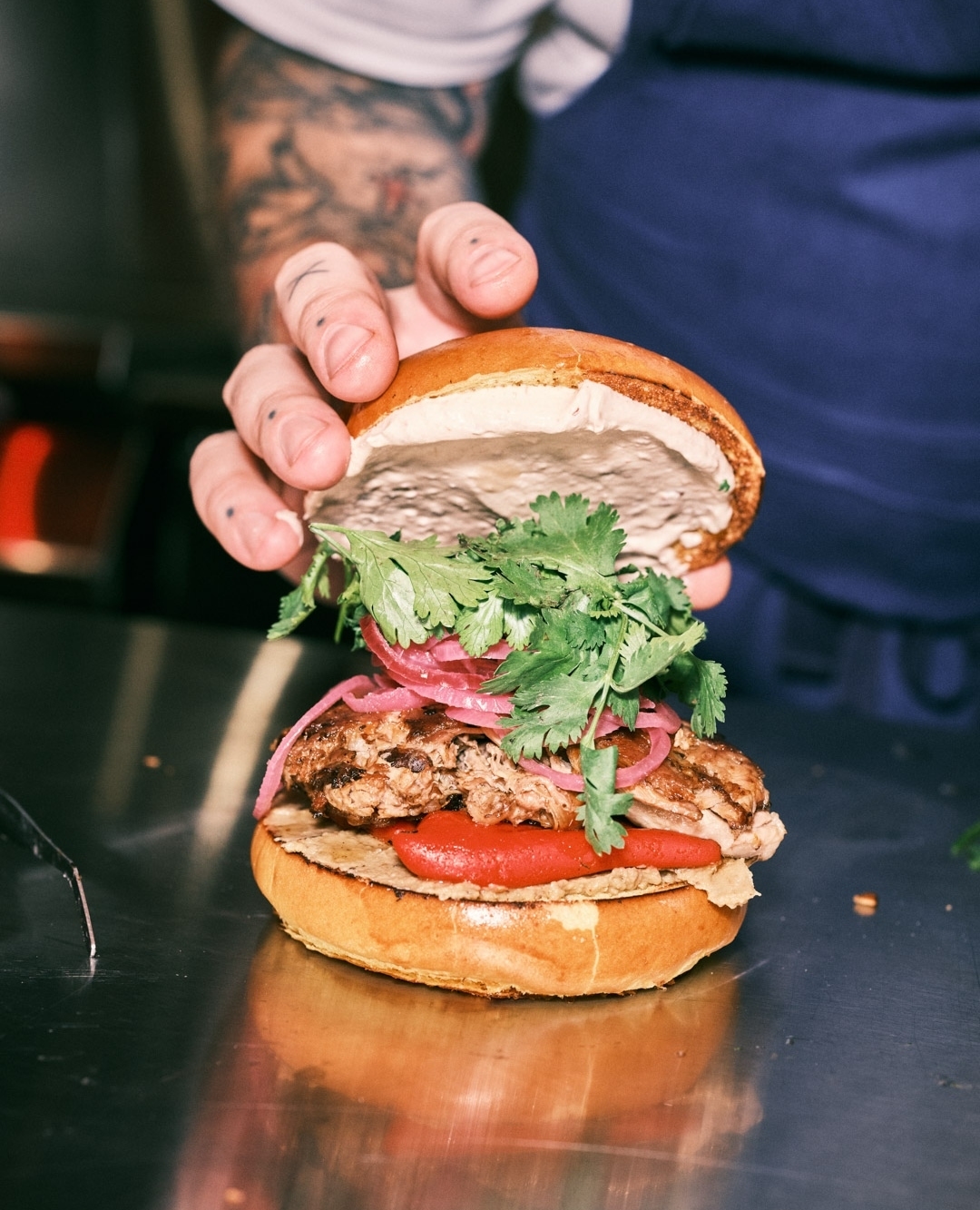 Close-up of a gourmet burger held by a hand with tattoos, featuring cilantro, red onion, and meat on a metal surface.