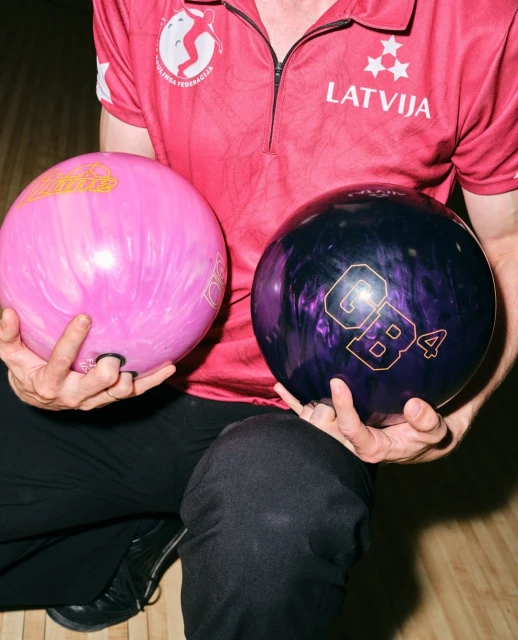 Man in pink uniform holds two bowling balls, one pink and one black, while kneeling. Close-up shot of sporting equipment indoors.