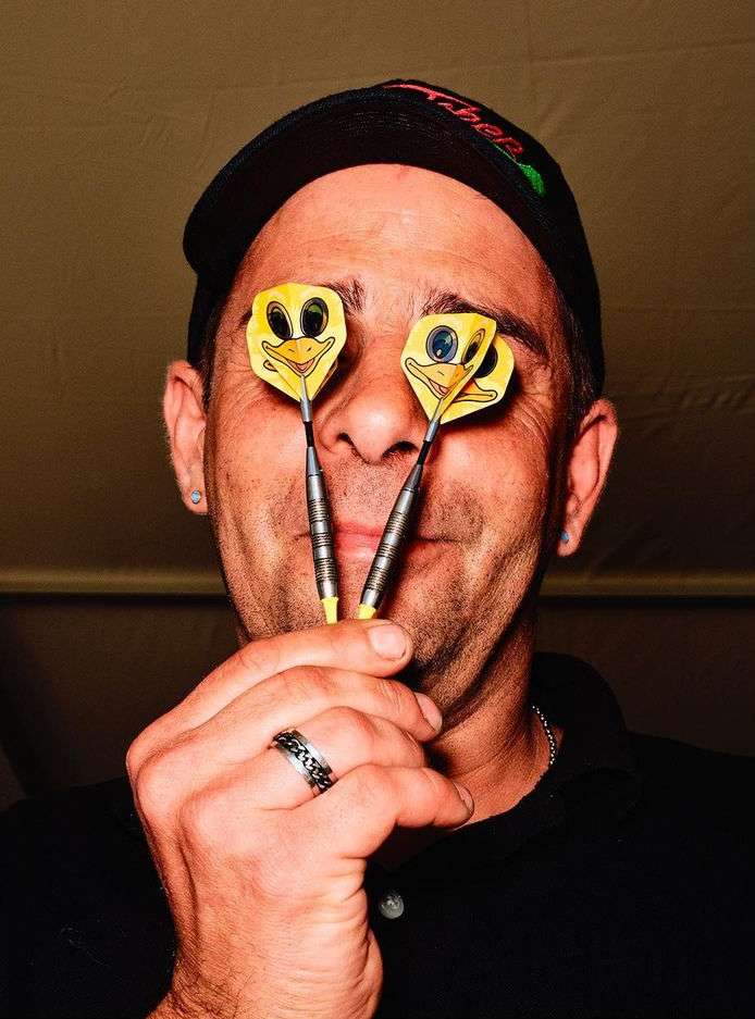 Adult male wearing a black cap and shirt, holding yellow dart arrows with cartoon duck faces up to his eyes and smiling.