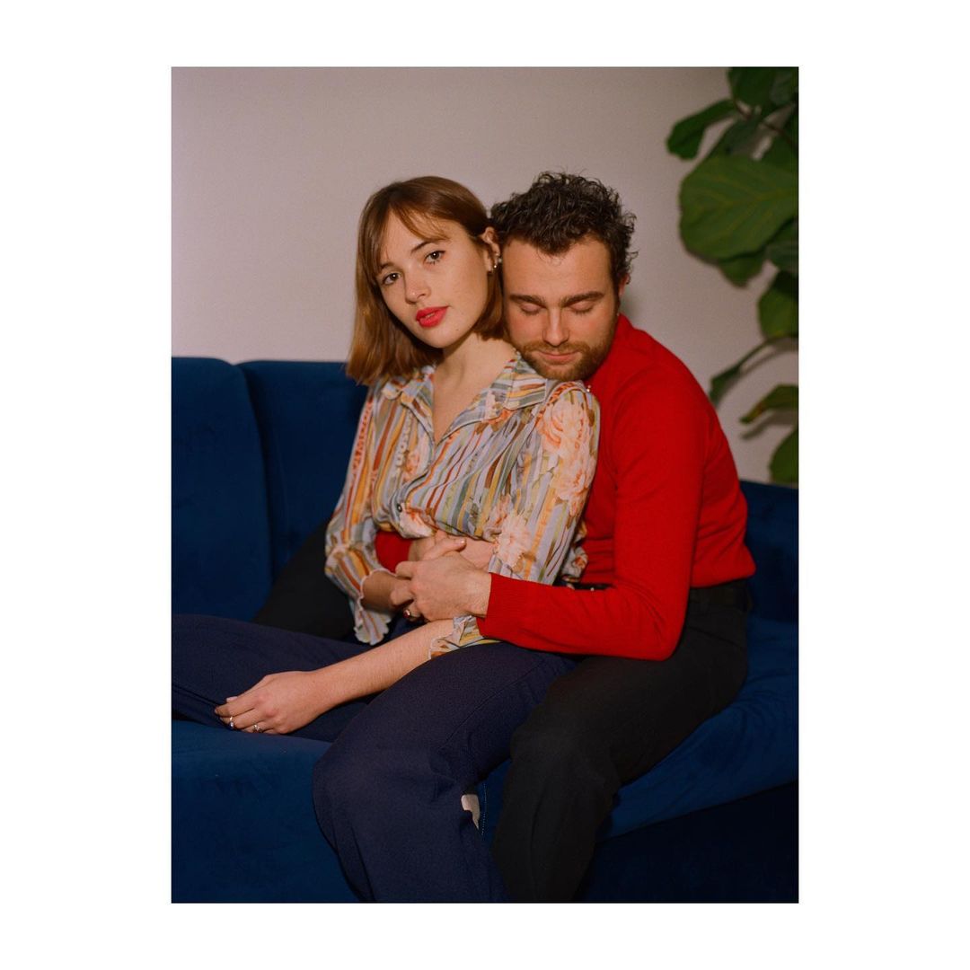 A couple embraces closely on a blue couch indoors, the woman in a patterned blouse and the man in a red sweater, with a potted plant nearby.