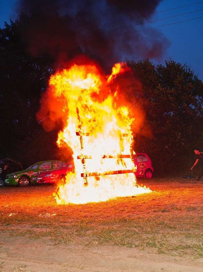 Large bonfire burning outdoors at night with cars in the background; fiery wood structure and flames.