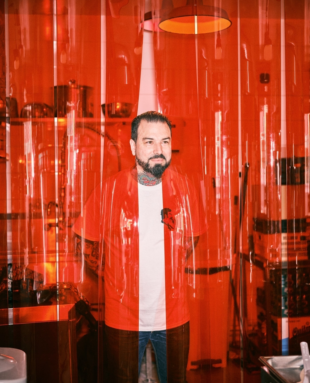 Man with beard and tattoos stands behind transparent plastic curtain indoors, wearing a white t-shirt and unbuttoned red overshirt.