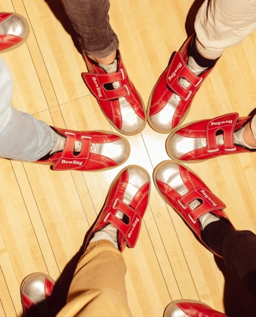 Overhead shot of six people wearing red and silver bowling shoes standing in a circle on a wood bowling alley floor.