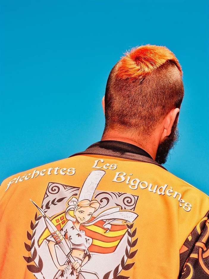 Adult male with orange mohawk and beard, wearing a yellow jacket with a female character and French text against a bright blue background.