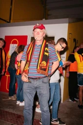Elderly man wearing a red cap, striped shirt, and football scarf smoking a cigarette indoors in Paris by Guillaume Blot