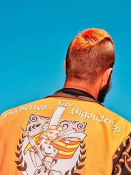 Rear view of a person with orange-dyed hair wearing an embroidered yellow jacket against a bright blue background