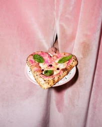Heart-shaped pizza with basil leaves on a white plate held against pink curtains by Guillaume Blot in Paris