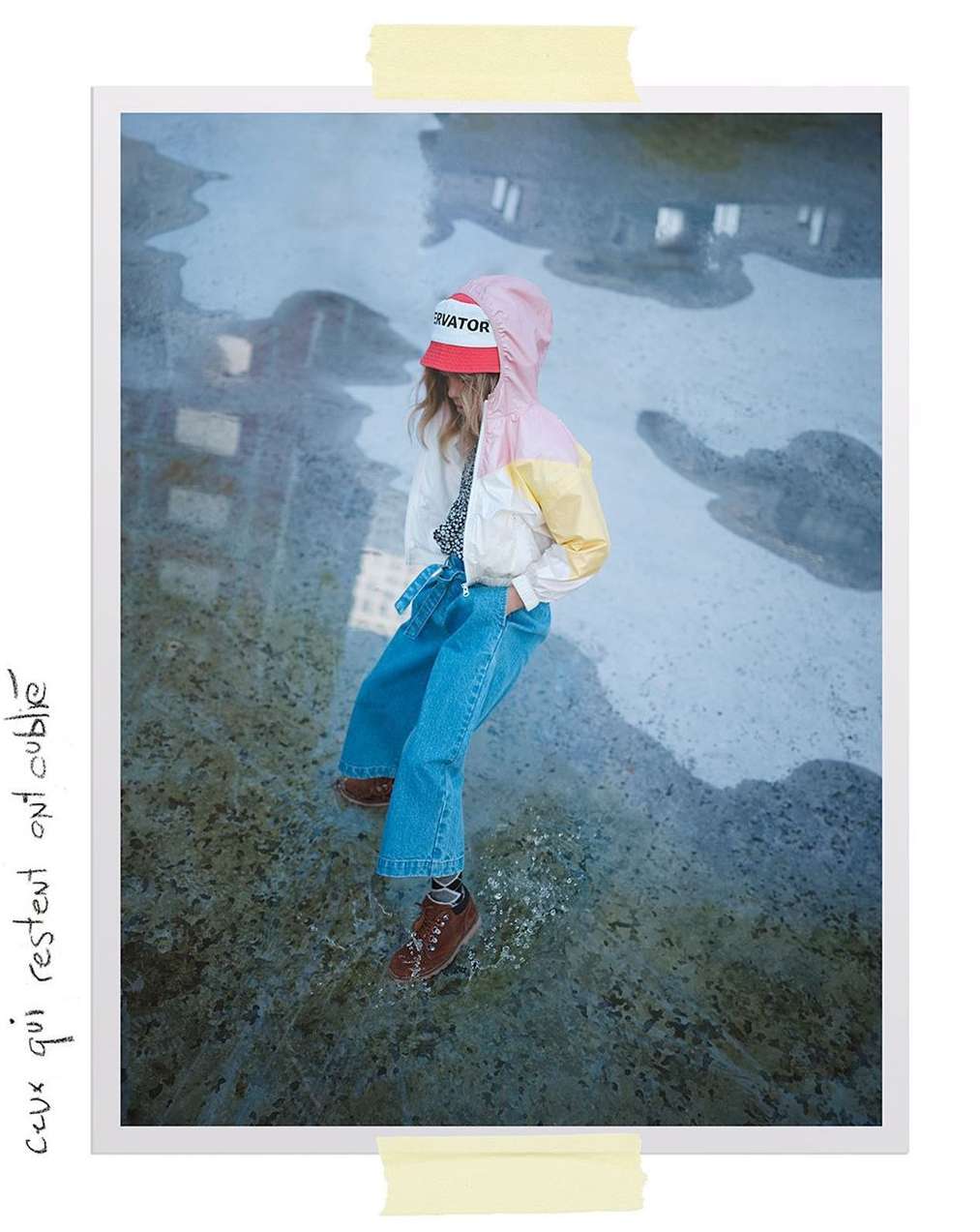 Child in a hooded color-blocked jacket, red cap, and wide-leg jeans stands in a puddle with city reflections in a Polaroid-style image.