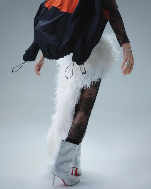 Fashion-forward studio shot featuring an adult female model wearing a black and orange jacket, white feather skirt, black lace tights, and silver boots.