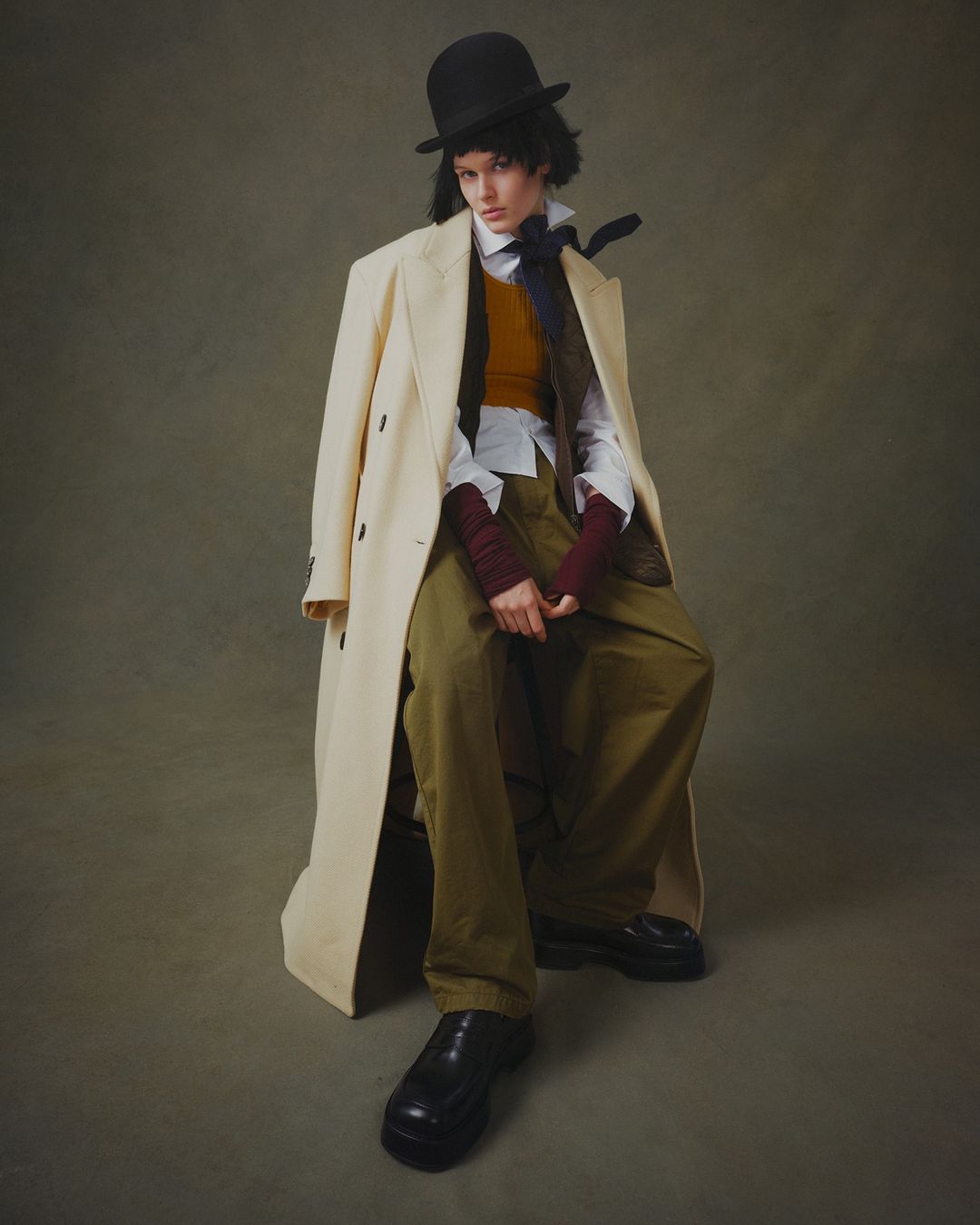 A young adult woman sits while modeling a bowler hat, tan overcoat, button-down shirt, orange vest, and green pants in a studio setting.