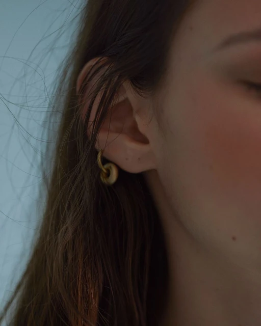 Close-up of a woman's ear with a gold hoop earring, dark hair, fair skin, and a blurred background.