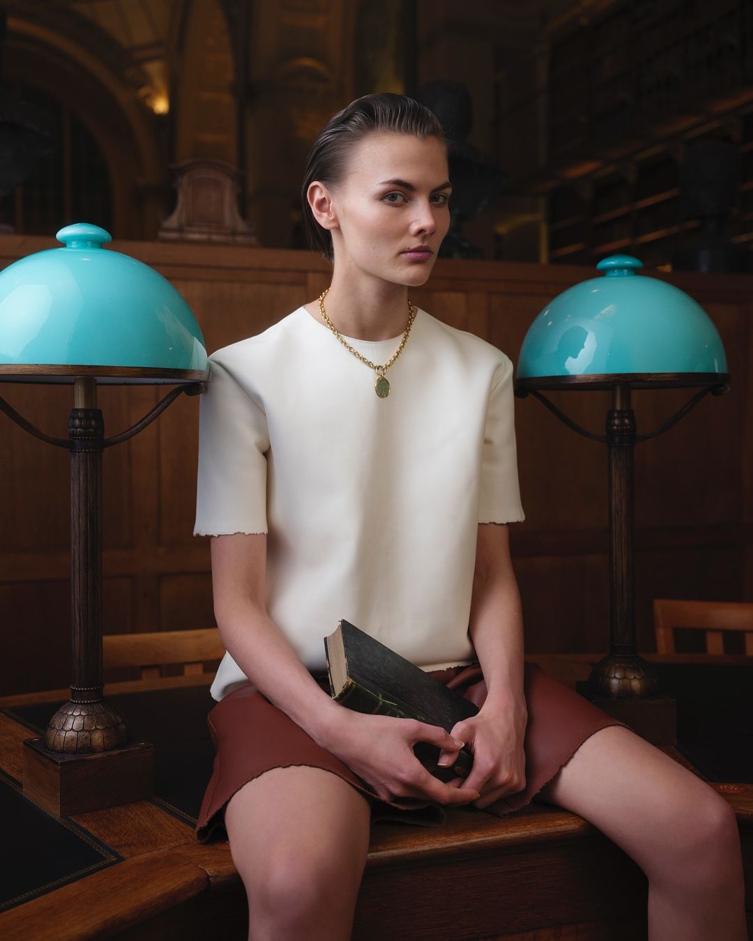Woman in white top and brown skirt sits on desk with lamps in library, holding book. Fashion shot with library background.