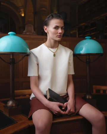 Woman in white top and brown skirt sits on desk with lamps in library, holding book. Fashion shot with library background.