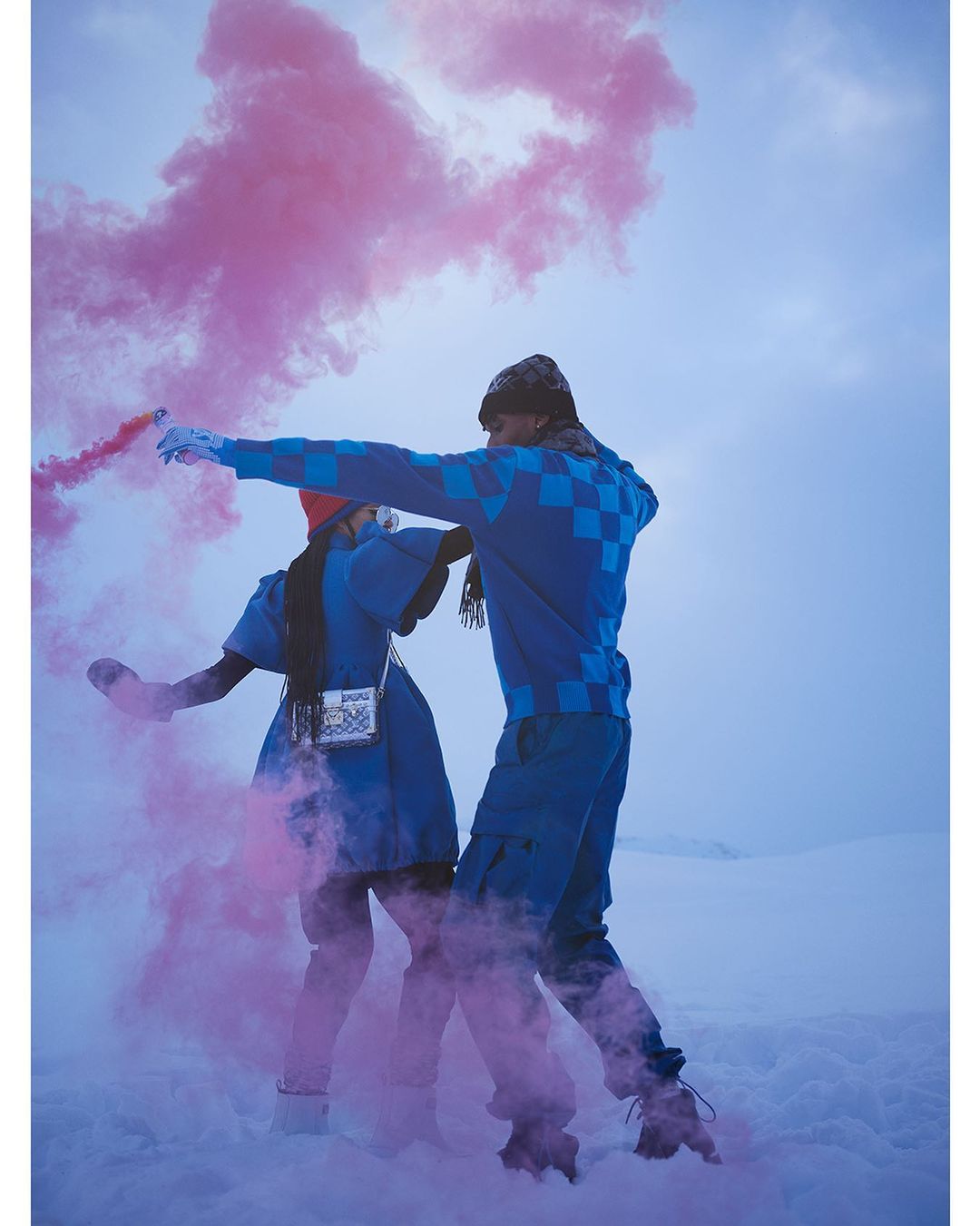Two people, a man and a woman, are standing in a snowy field with pink smoke around them. Both are wearing blue outfits.