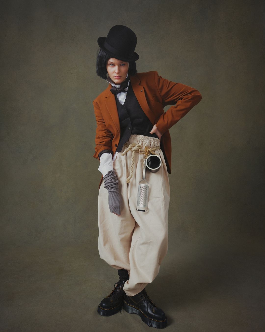 Full-length portrait of a woman in a bowler hat, orange blazer, vest, and beige pants, posing in front of a dark green backdrop.