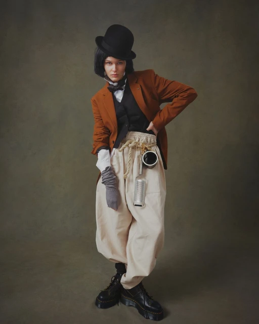 Full-length portrait of a woman in a bowler hat, orange blazer, vest, and beige pants, posing in front of a dark green backdrop.