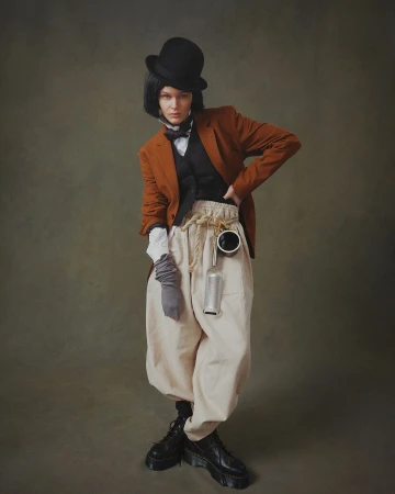 Full-length portrait of a woman in a bowler hat, orange blazer, vest, and beige pants, posing in front of a dark green backdrop.