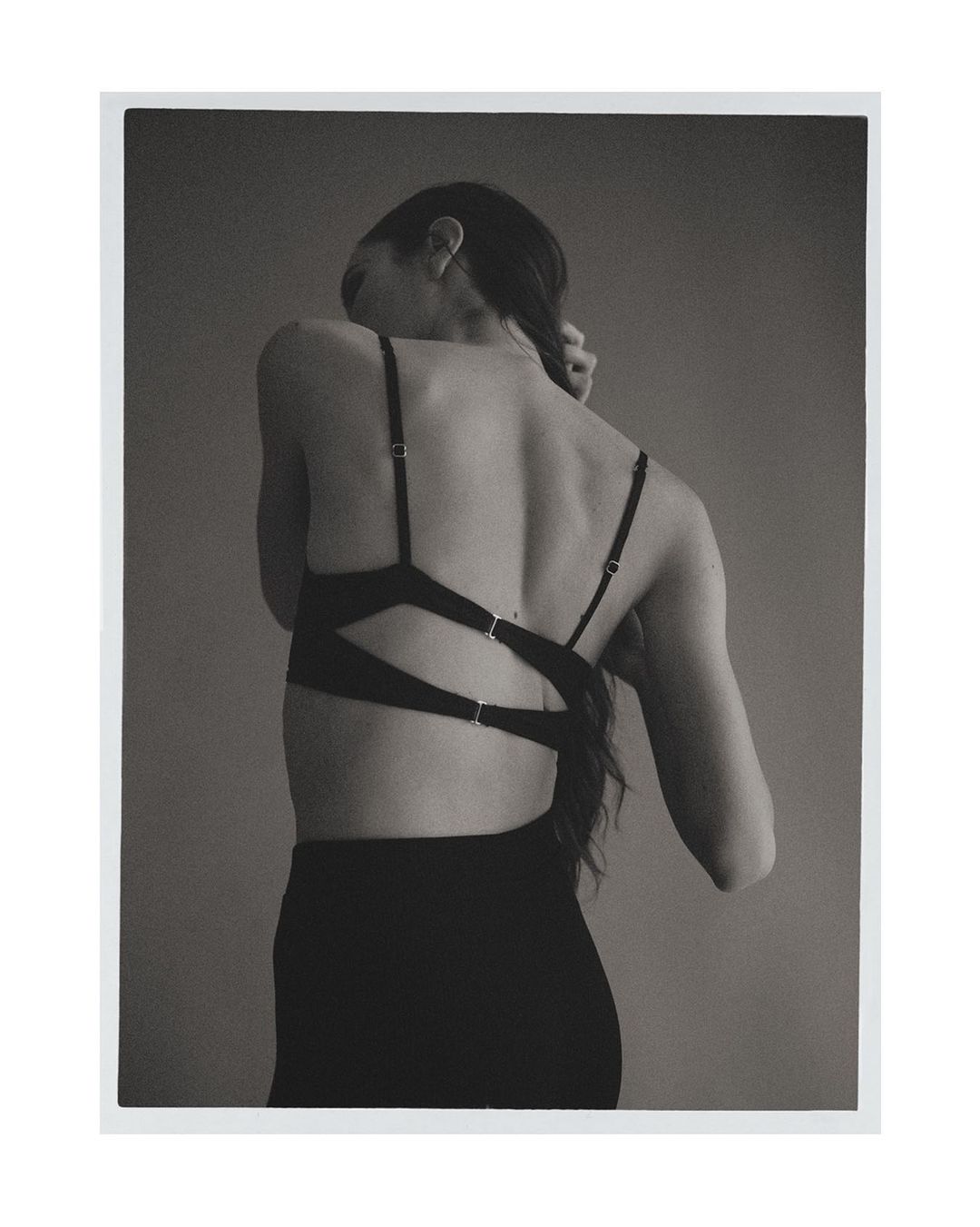 Back view of a woman with braided hair wearing a black bralette and skirt against a neutral gray background in a minimalist style.