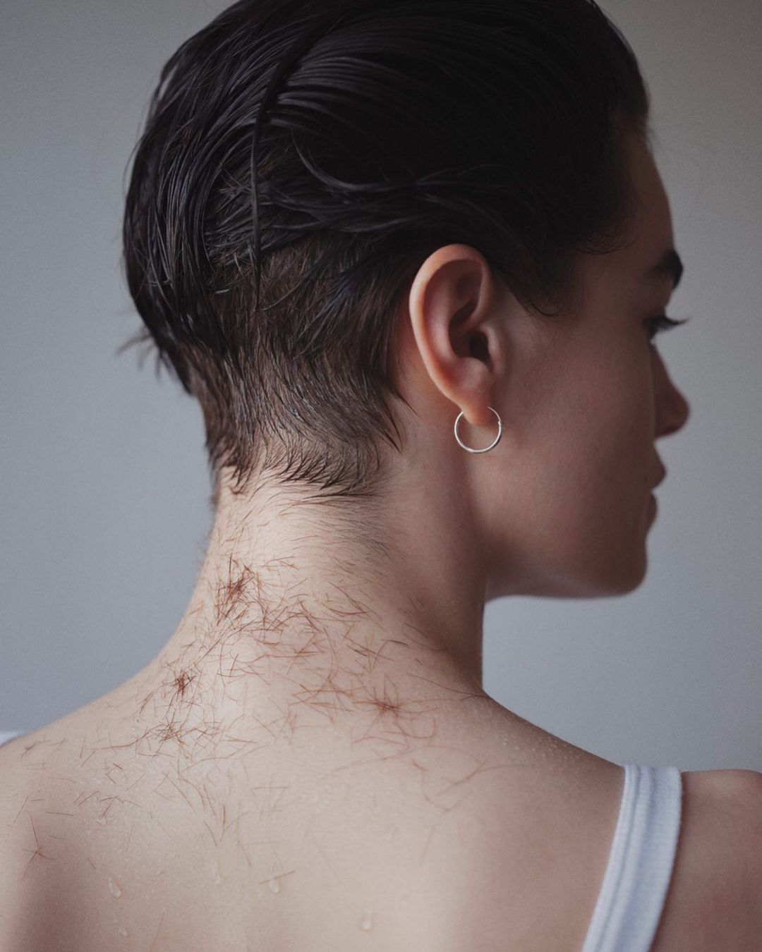 Portrait of person with shaved neck and short wet hair. Shoulder and back skin with hair clippings visible against soft grey background.
