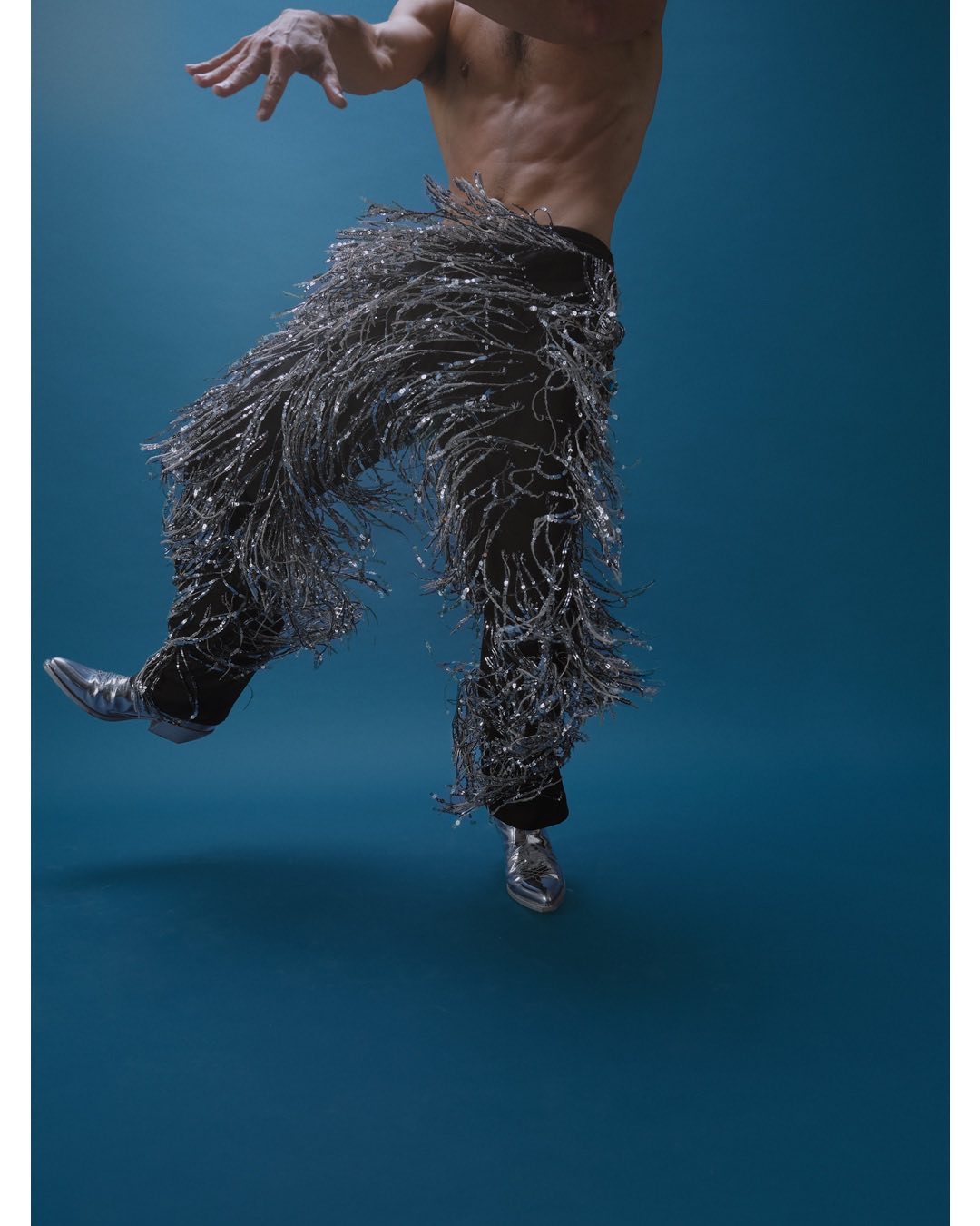 Shirtless man dancing in silver fringed pants and metallic boots against a dark blue background