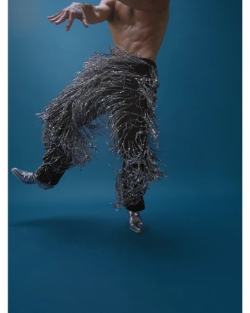 Shirtless man dancing in silver fringed pants and metallic boots against a dark blue background