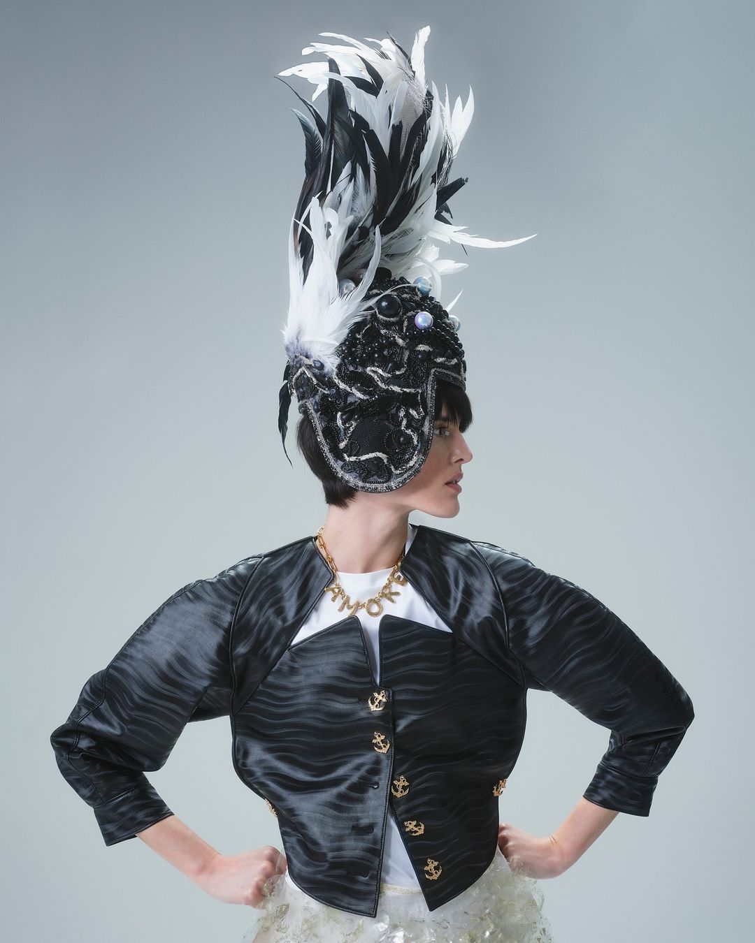 Adult female model posing in a black-and-white textured jacket, gold anchor buttons, gold necklace, and large feathered headpiece in studio.
