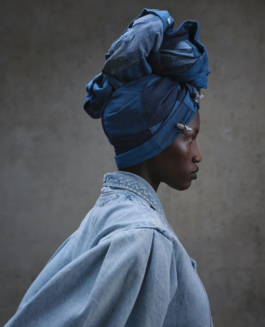 Profile portrait of a dark-skinned woman wearing a denim headwrap and jacket against a textured grey background in a photo studio.
