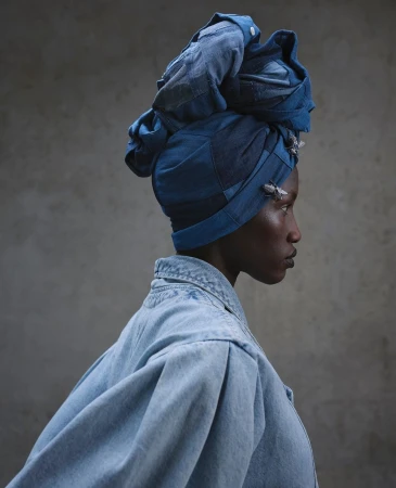 Profile portrait of a dark-skinned woman wearing a denim headwrap and jacket against a textured grey background in a photo studio.