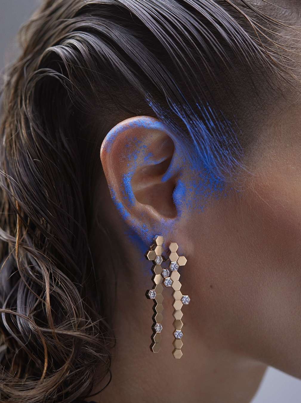 Close-up of a woman with wavy brown hair and blue makeup around her ear wearing gold-tone drop earrings with clear gems.