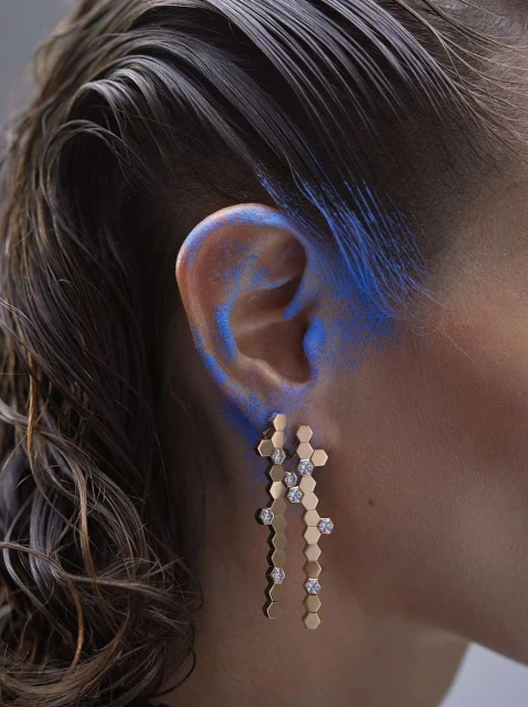 Close-up of a woman with wavy brown hair and blue makeup around her ear wearing gold-tone drop earrings with clear gems.