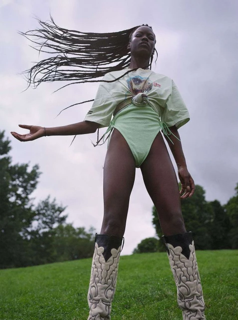 Low-angle shot of a Black woman in a pale green swimsuit and cowboy boots. Wind blows through her hair on a grassy field.