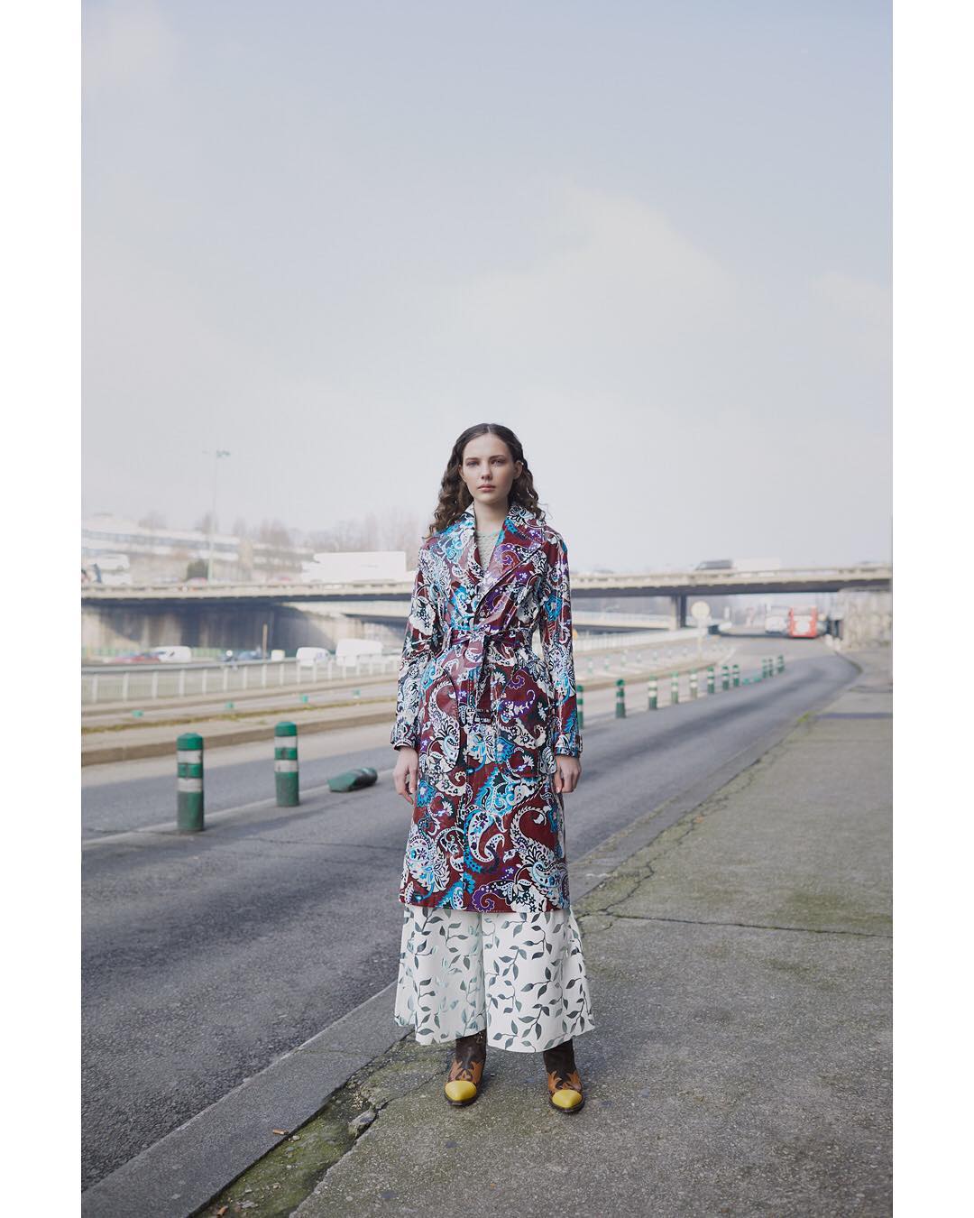 A woman in a patterned trench coat and floral pants poses on an urban street, showcasing a blend of fashion and urban style in daylight.