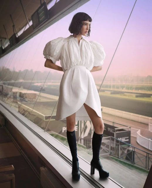 Fashionable woman in white dress and boots posing inside a glass enclosure with a racetrack view, capturing a blend of style and location.