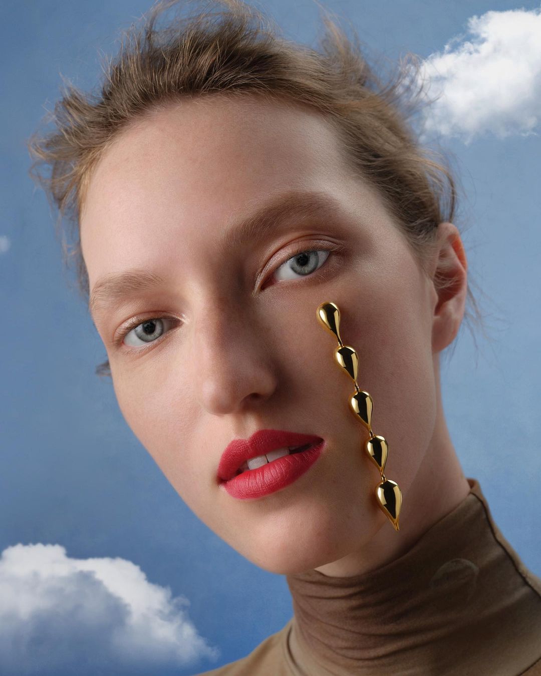 Close-up portrait of a model with red lipstick and a chain of gold teardrop-shaped jewelry hanging from her eye against a cloudy sky.