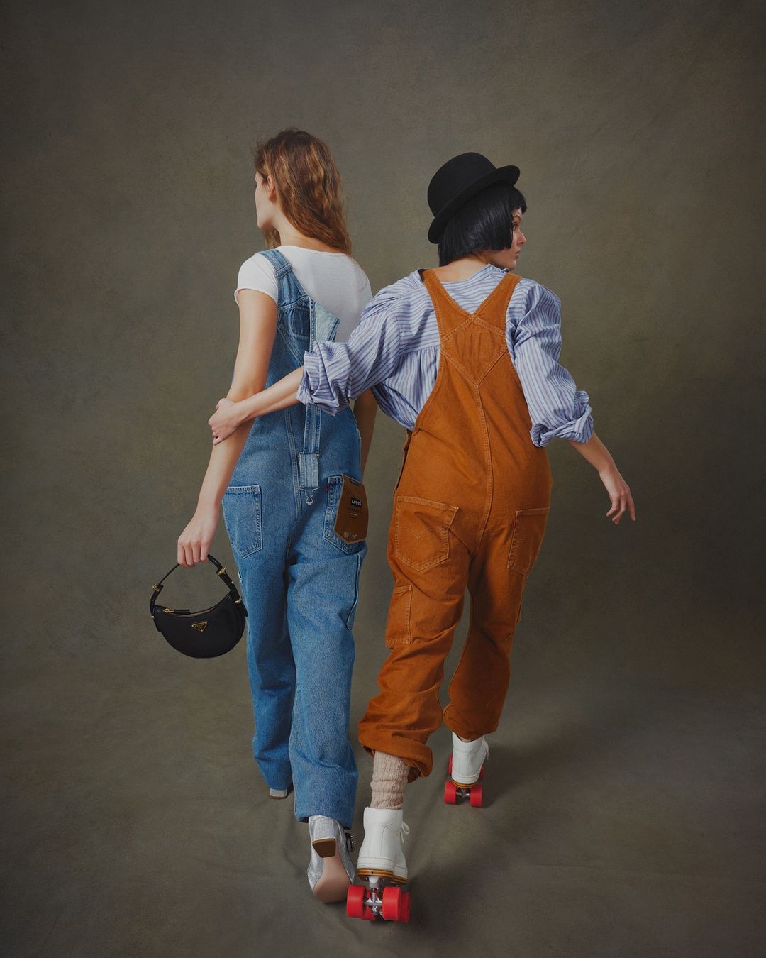 Two people on roller skates, one in blue overalls carrying a purse, the other in brown overalls and a bowler hat, walking away.