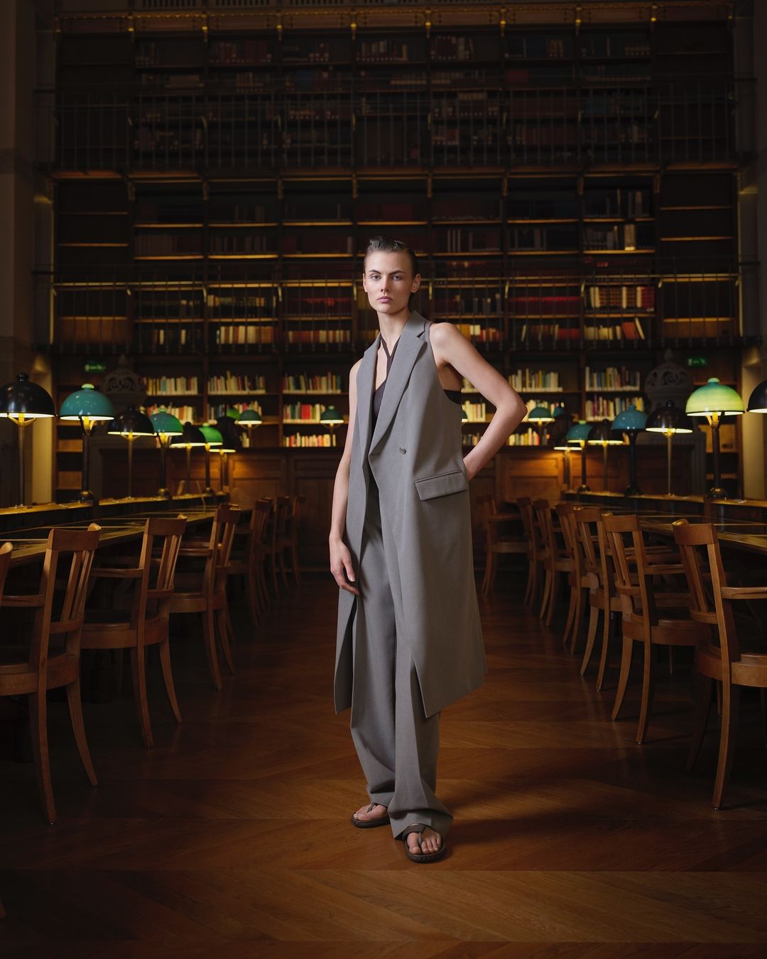 Fashion shot in a library: a woman in a long grey vest and pants stands in the center of a library with rows of books.
