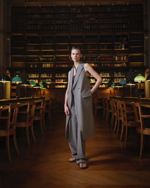 Fashion shot in a library: a woman in a long grey vest and pants stands in the center of a library with rows of books.