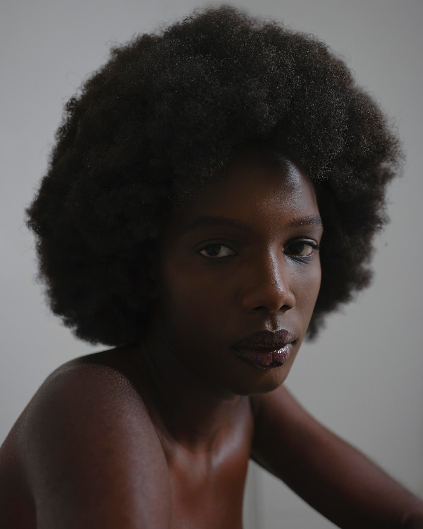 Close-up portrait of a Black woman with voluminous afro hair and glossy dark lips against a plain grey backdrop