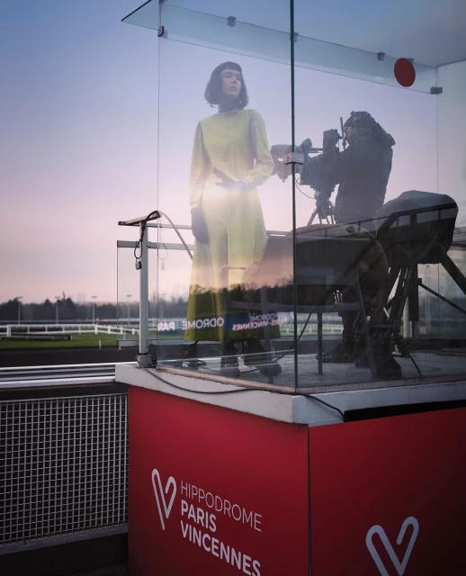 Woman in a yellow outfit stands inside a glass booth as a cameraman films her at the Hippodrome Paris Vincennes racecourse.