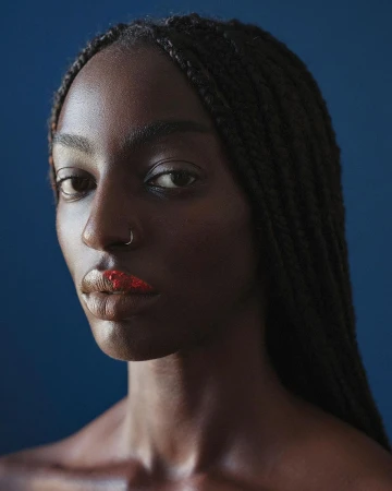 Close-up portrait of a striking, dark-skinned woman with braids, wearing red lipstick and a nose ring against a dark blue backdrop.